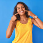 Brown woman in a yellow tank top smiles after professional teeth whitening while standing against a blue wall
