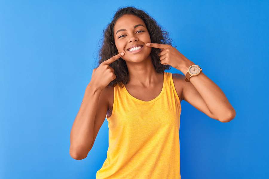 Brown woman in a yellow tank top smiles after professional teeth whitening while standing against a blue wall