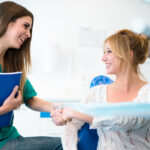 A blonde patient in a dental chair smiles as her dental hygienist greets her at the dentist