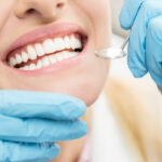 Closeup of a woman smiling at the dentist during her routine cleaning as blue gloved hands hold a special dental mirror