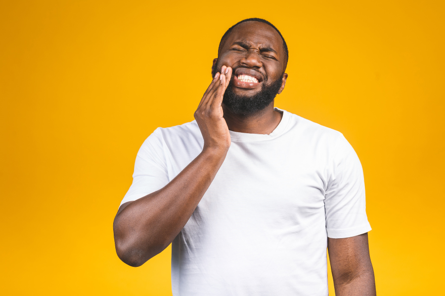 Black man in a white t-shirt cringes in pain and touches his cheek due to teeth sensitivity