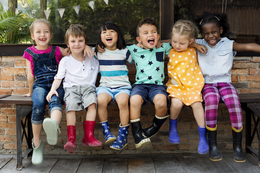 A group of young children smile as the sit on a bench together while wearing rain boots