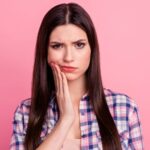 Brunette woman in a plaid shirt cringes and touches her cheek in pain due to sensitivity against a pink background