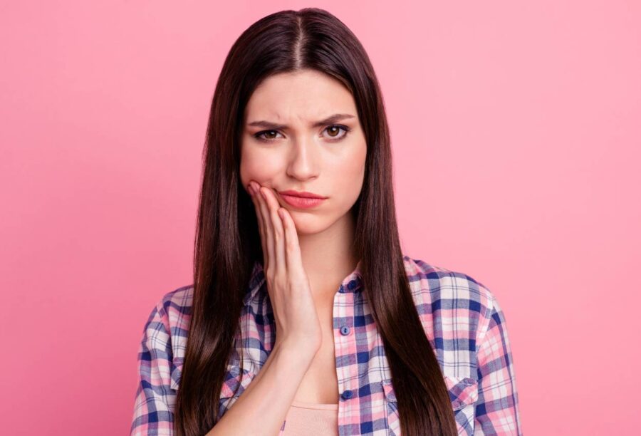 Brunette woman in a plaid shirt cringes and touches her cheek in pain due to sensitivity against a pink background