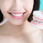 A young woman holds up clear aligners next to a teeth model wearing traditional braces