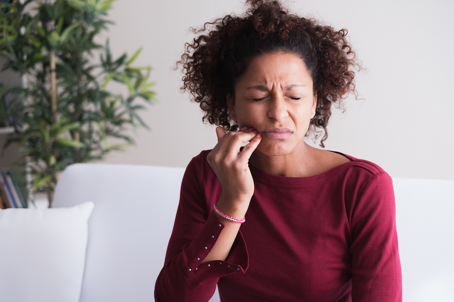 Black woman in a burgundy sweater cringes in pain and touches her cheek due to a toothache